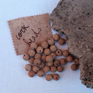 以下が含まれることがあります： 茶色のコルクビーズの山をクローズアップした写真。各ビーズには小さな穴が開いています。ビーズは白い布の表面に置かれており、その上に「コルクビーズ」と黒インクで書かれたコルク生地の切れ端があります。