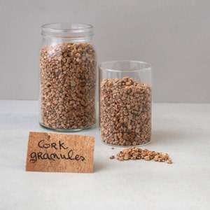 May include: Two clear glass jars filled with brown cork granules. One jar is a wide-mouth mason jar, and the other is a cylindrical glass. A small piece of cork with the words "Cork granules" is in front of the jars.