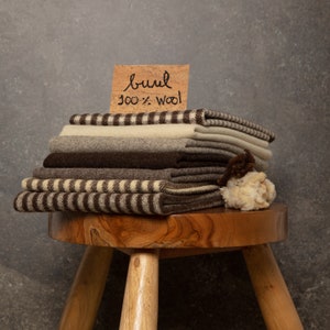 May include: A stack of folded wool fabric pieces in shades of brown, beige, and cream, featuring solid and striped patterns. A rectangular cork sign rests on top with the handwritten text "burel 100% wool". A small tuft of raw wool sits beside the stack.