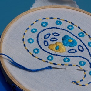 May include: A white embroidery hoop with a blue and yellow paisley design. The design features a paisley shape with blue and yellow floral accents.