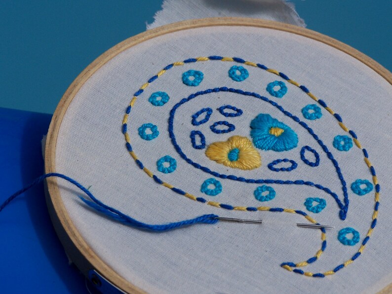 May include: A white embroidery hoop with a blue and yellow paisley design. The design features a paisley shape with blue and yellow floral accents.