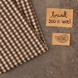 May include: A piece of fabric with a brown and cream houndstooth pattern. Next to the fabric are two rectangular cork labels. One label reads "burel 100% wool" in black ink, and the other features a paisley design with the text "FOLCLORE".
