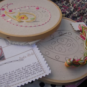 May include: Two embroidery hoops with a paisley design. The top hoop shows a completed embroidery with pink, yellow, and green thread. The bottom hoop shows a blank paisley design with a skein of pink, green, and white thread. The text on the paper reads "Ponto Atr&aacute;s".