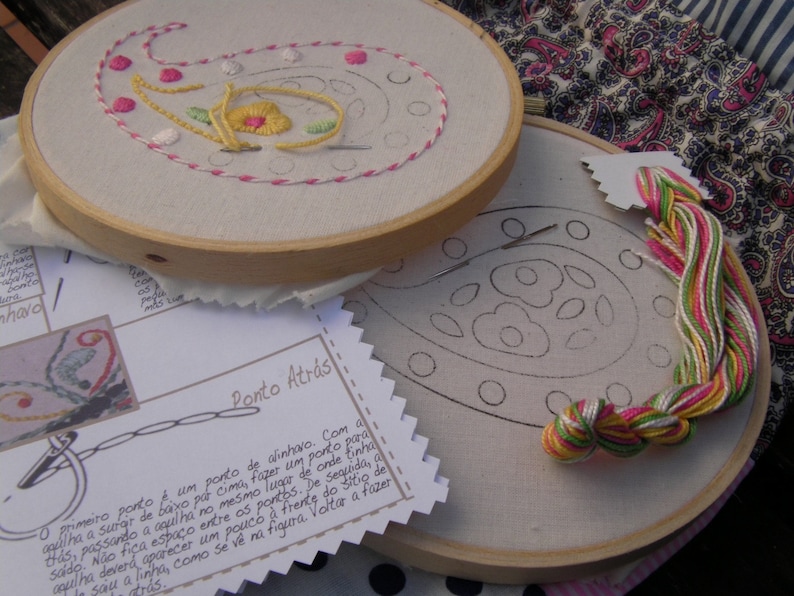 May include: Two embroidery hoops with a paisley design. The top hoop shows a completed embroidery with pink, yellow, and green thread. The bottom hoop shows a blank paisley design with a skein of pink, green, and white thread. The text on the paper reads "Ponto Atr&aacute;s".