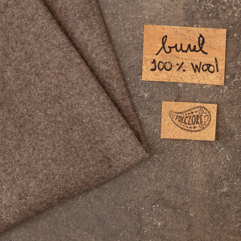 May include: A folded piece of brown felted fabric with a textured surface. Next to the fabric are two rectangular cork labels. One label has the handwritten text "burel 100% wool" and the other features a paisley design with the word "FOLCLORE".