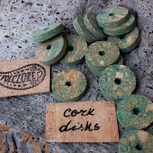 Op de afbeelding: Een verzameling van 15 groene en bruine kurk schijven met een gat in het midden. De schijven zijn op een getextureerde ondergrond in een stapel gelegd. Een klein kurk label met het woord "FOLKLORE" is zichtbaar in de afbeelding. Een tweede kurk label met de tekst "Kork schijven" is ook zichtbaar.