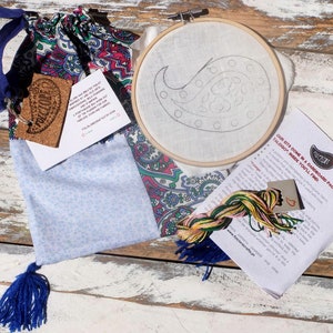 May include: A paisley embroidery kit containing a blue and white fabric bag, a wooden embroidery hoop, embroidery floss, and instructions. The kit features a pre-printed design of a paisley pattern on the fabric.