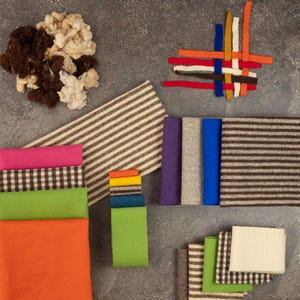 May include: A collection of textile materials including loose brown and cream wool fibers, various solid-colored fabric swatches in pink, green, orange, purple, gray, and blue, and several pieces of fabric featuring brown and cream striped or checkered patterns.