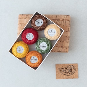 Puede incluir: Una caja blanca llena de seis carretes de hilo de bordar de varios colores, incluyendo rojo, verde, amarillo y marrón. Los carretes están etiquetados con "Limol Perle" y tienen un número en cada carrete. La caja está sobre una superficie de corcho con una pequeña etiqueta de corcho que dice "Folklore".