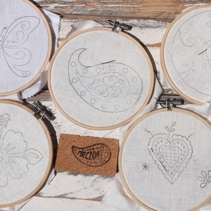 May include: Five embroidery hoops with different designs: a butterfly, a paisley, a flower, a heart, and a sun. Each hoop has a needle threaded through the fabric.