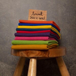 May include: A stack of folded wool fabric in various colors including orange, blue, yellow, teal, red, purple, pink, green, and mustard. A rectangular cork sign sits on top with the handwritten text "burel 100% wool" in black ink.