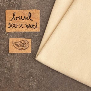 May include: A folded piece of cream-colored wool fabric next to two rectangular cork labels. The top label features the handwritten text "burel 100% wool" in black ink, while the bottom label displays a paisley-shaped logo with the word "FOLCLORE".