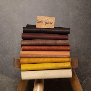 May include: A stack of cork fabric samples in various colors, including brown, red, orange, yellow, and white. The samples are arranged on a wooden stool with a note that reads "Cork Fabric".