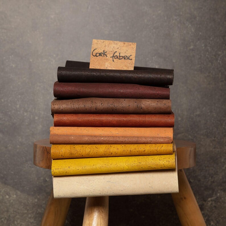 May include: A stack of cork fabric samples in various colors, including brown, red, orange, yellow, and white. The samples are arranged on a wooden stool with a note that reads "Cork Fabric".