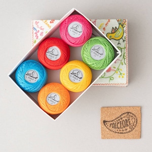 Può includere: Un set di sei rocchetti di filo da ricamo colorati in una scatola bianca. I rocchetti sono etichettati "Limol Perle" e sono di colore rosso, arancione, giallo, verde, blu e rosa.