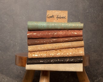 May include: A stack of cork fabric in various colors and patterns, including teal, brown, beige, and black. The fabric is stacked on a wooden stool with a sign that reads "Cork fabric".