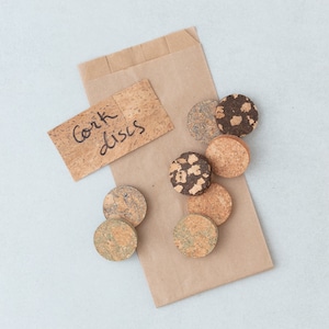 May include: A brown paper bag with a label that says "Cork discs". The bag contains a variety of cork discs in different patterns and colours.