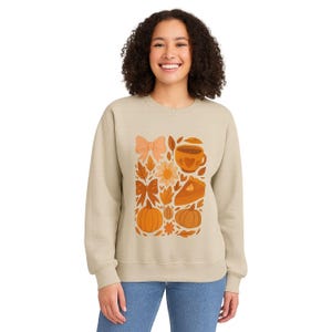 May include: Beige sweatshirt with a fall-themed graphic. The design features a bow, flowers, pumpkins, a coffee cup, and a slice of pie in orange and brown hues. The sweatshirt is a casual, comfortable style.