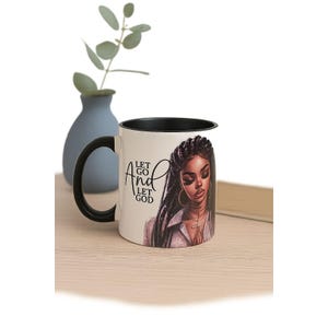 May include: A white ceramic mug with a black handle and rim. The mug features an illustration of a woman with long braids and the text "Let Go And Let God". A blue vase with a plant is in the background.