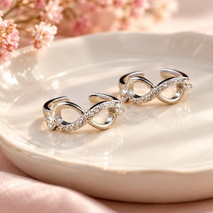 May include: Two silver-toned open rings featuring an infinity symbol design. Each ring is accented with small, clear, sparkling stones embedded along the infinity loop, creating a continuous, reflective surface on the metallic band structure.