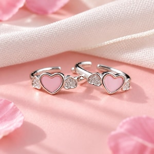 Puede incluir: Un par de pendientes de aro plateados con un corazón rosa central flanqueado por dos pequeños corazones incrustados con piedras transparentes brillantes. El diseño es simétrico, con un acabado metálico pulido y un cierre de bisagra.