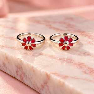 以下が含まれることがあります： 鮮やかな赤色のエナメル花びらと中央の銀色の金属ドットが特徴の、花のデザインが施された2つの銀色の指輪。指輪はオープンバンド構造になっており、さまざまな指のサイズに合わせて柔軟に調整することが可能です。