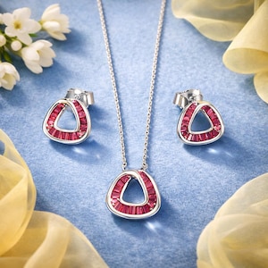 Peut inclure: Un ensemble de bijoux en argent comprenant un collier et des boucles d'oreilles. Le pendentif et les boucles d'oreilles présentent un design triangulaire orné de pierres précieuses rouges. L'ensemble est présenté sur un fond bleu clair.