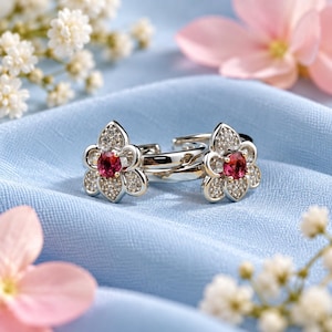 May include: A pair of silver-toned hoop earrings featuring a flower-shaped design. Each earring is centered with a round, deep pink gemstone and surrounded by small, clear pavé-set stones on the petals, creating a bright and reflective finish.