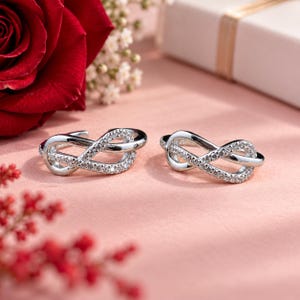 Peut inclure: Une paire de boucles d'oreilles en forme de symbole infini argenté, dotées d'une finition métallique polie et d'une rangée de petites pierres claires et scintillantes incrustées le long de la courbe avant, formant une boucle continue.