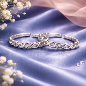 Puede incluir: Dos pulseras plateadas de extremo abierto con un diseño de símbolo de infinito. Las pulseras se exhiben sobre una tela de satén azul, con flores blancas y una cinta rosa en el fondo. Las pulseras probablemente están hechas de plata de ley.