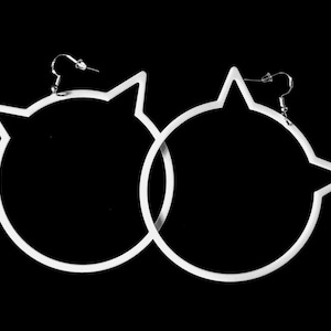 Puede incluir: Un par de pendientes de aro blancos con forma de gato. Los pendientes presentan un diseño circular con orejas puntiagudas en la parte superior. Los pendientes están suspendidos de ganchos plateados sobre un fondo negro.