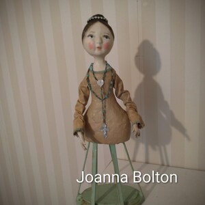 May include: A handmade doll with a serene expression, wearing a tan dress and a beaded necklace with a cross pendant. The doll is mounted on a green metal stand. The text "Joanna Bolton" is visible at the bottom.