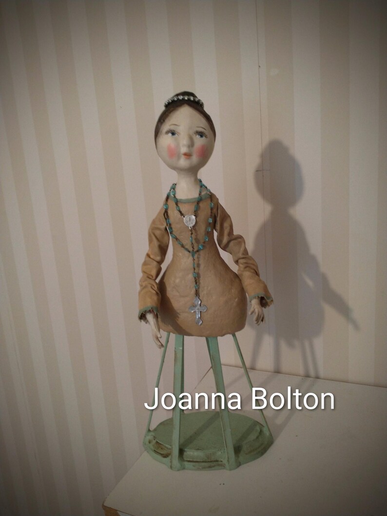 May include: A handmade doll with a serene expression, wearing a tan dress and a beaded necklace with a cross pendant. The doll is mounted on a green metal stand. The text "Joanna Bolton" is visible at the bottom.