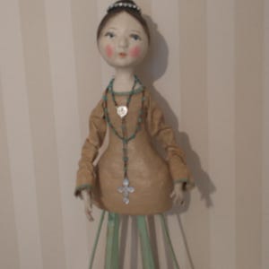 May include: A handcrafted doll with a serene expression, wearing a tan dress and a turquoise beaded necklace with a cross pendant. The doll has a delicate tiara and is mounted on a green metal stand.