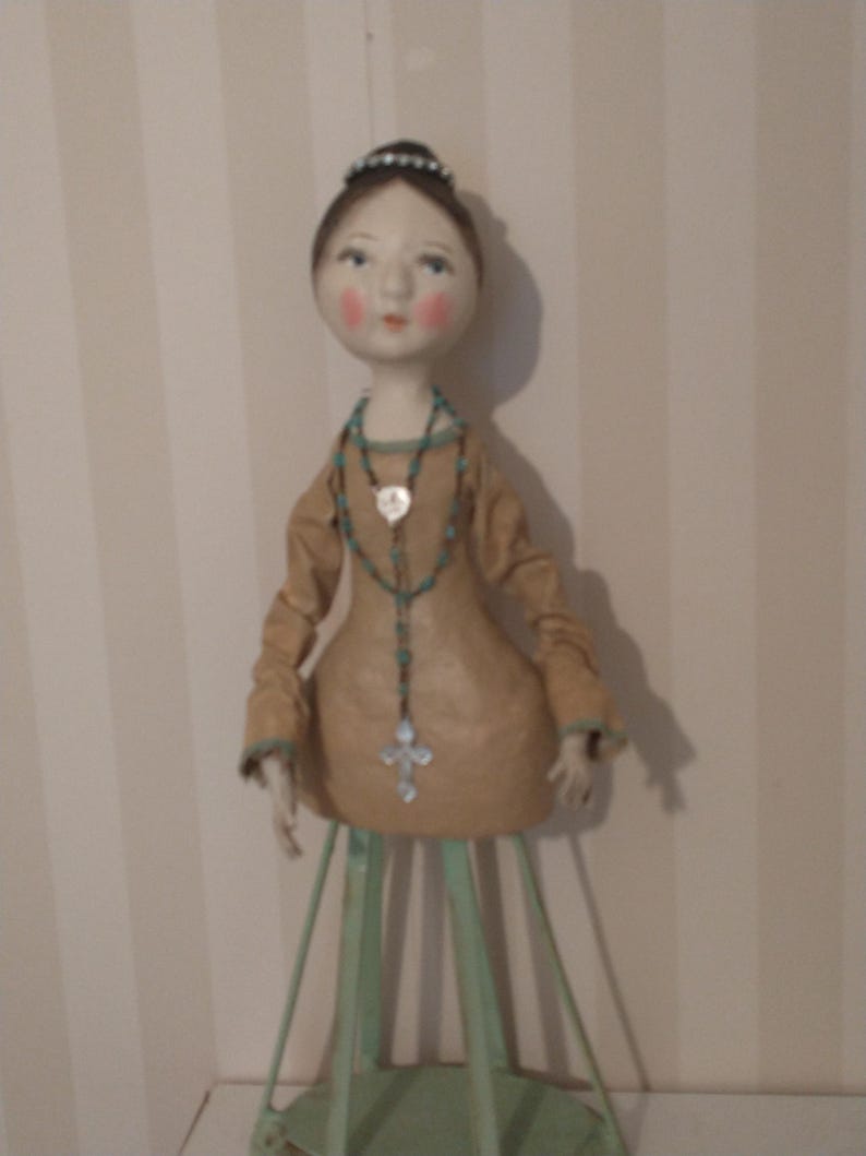 May include: A handcrafted doll with a serene expression, wearing a tan dress and a turquoise beaded necklace with a cross pendant. The doll has a delicate tiara and is mounted on a green metal stand.