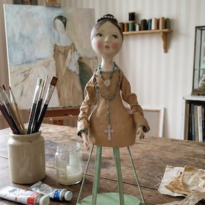 A 16-inch handcrafted OOAK Santos cage doll in a Modern Primitive folk art style. The mixed-media sculpture features a hand-sculpted clay head and torso with a serene expression, attached to a wooden slat 'cage' base. The doll is presented in an artist's studio with signature striped wallpaper, creating a nostalgic, antique feel