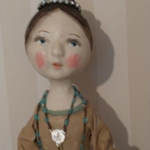 May include: A handmade doll with a serene expression. The doll has a pale face with rosy cheeks, blue eyes, and a small tiara. It wears a tan dress and a turquoise beaded necklace with a silver pendant. The doll's hair is styled in a bun.