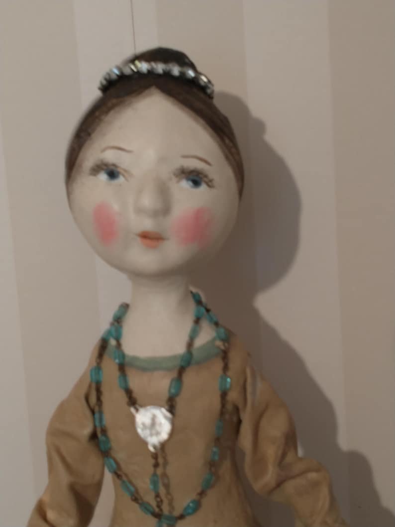May include: A handmade doll with a serene expression. The doll has a pale face with rosy cheeks, blue eyes, and a small tiara. It wears a tan dress and a turquoise beaded necklace with a silver pendant. The doll's hair is styled in a bun.