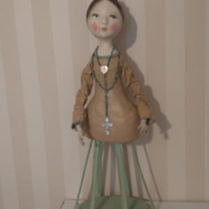 May include: A handcrafted doll with a tan dress and a turquoise beaded necklace with a cross pendant. The doll has a delicate face with rosy cheeks and a small tiara. It stands on a green metal stand.