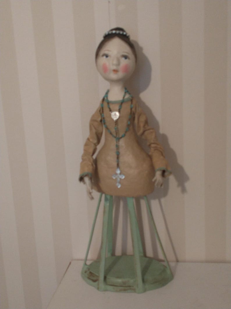 May include: A handcrafted doll with a tan dress and a turquoise beaded necklace with a cross pendant. The doll has a delicate face with rosy cheeks and a small tiara. It stands on a green metal stand.