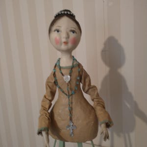 May include: A handcrafted doll with a serene expression, wearing a tan dress with turquoise trim. The doll has a turquoise beaded necklace with a silver cross pendant. The doll's hair is styled in a bun, and it is mounted on a green stand.