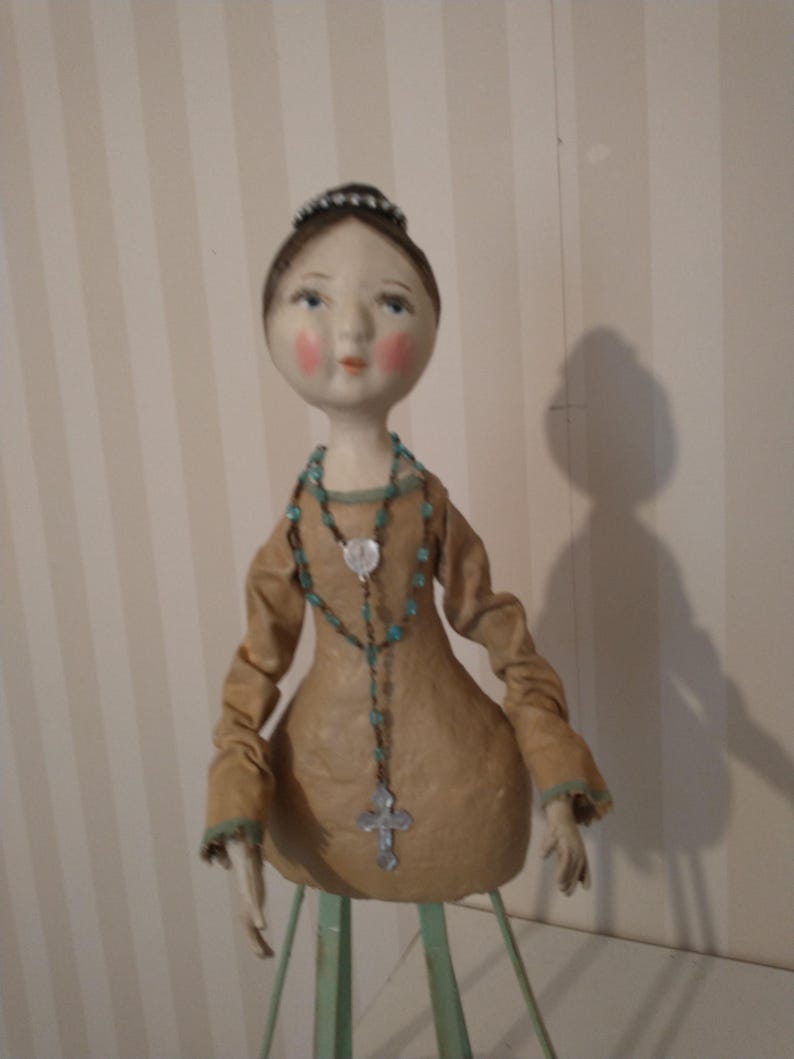 May include: A handcrafted doll with a serene expression, wearing a tan dress with turquoise trim. The doll has a turquoise beaded necklace with a silver cross pendant. The doll's hair is styled in a bun, and it is mounted on a green stand.