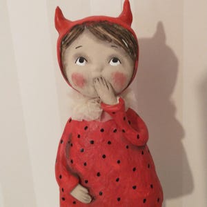 May include: A figurine of a child wearing a red hood with two small horns, a red dress with black polka dots, and a white ruffled collar. The figure has brown hair, rosy cheeks, and one hand raised to cover their mouth in a gesture of surprise.