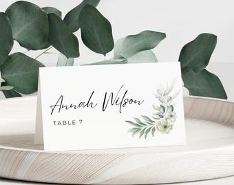 May include: White place card with green and white floral design. The text on the card reads "Anna Wilson, Table 7".