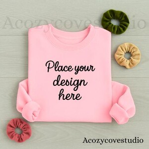 May include: A light pink sweatshirt with the text "Place your design here" in black script. Three scrunchies in green, tan, and red are arranged around the sweatshirt. The background is a light gray wood grain.