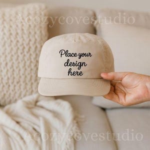 Puede incluir: Gorra de béisbol beige con el texto "Place your design here" en escritura negra. La gorra está sujeta por una mano, con un fondo de tonos suaves y neutros.