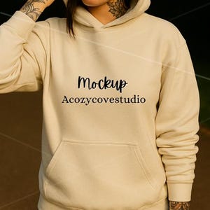 May include: A cream-coloured hoodie with a front pocket and a drawstring hood. The word "Mockup" is printed in black script above the text "Acozycovestudio". The hoodie has long sleeves and ribbed cuffs.