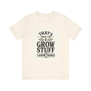 May include: A cream-colored short-sleeve t-shirt featuring dark green text that reads: THAT'S WHAT I DO I GROW STUFF AND I KNOW THINGS. The design includes small leaf illustrations above and below the text, centered on the front of the shirt.