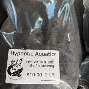 May include: A transparent plastic bag containing dark brown terrarium soil. The label states "Hypnotic Aquatics Terrarium soil Self sustaining" and indicates a weight of 0.9 kg.