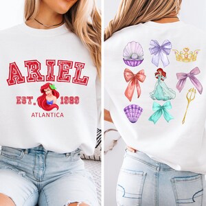 Disney Ariel sweatshirt in pastel blue with bold red varsity lettering on the front, reading 'Ariel Est. 1989 Atlantica.' The back design includes princess elements like seashells, bows, a golden trident, and an elegant illustration of